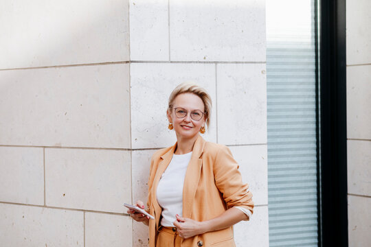 Smiling female entrepreneur with smart phone while standing against wall