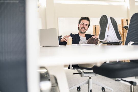 Young businessman sitting in office, with feet on desk, drinking coffee