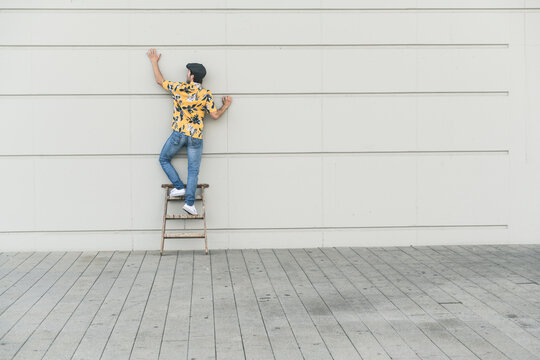 Young man wearing flat hat and aloa shirt, standing on step ladder, touching wall