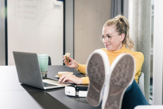 Smiling casual businesswoman sitting at table in office using laptop and eating a sandwich