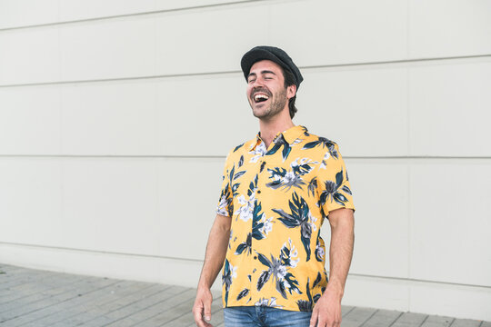 Young man wearing flat hat and aloha shirt, laughing in front of wall