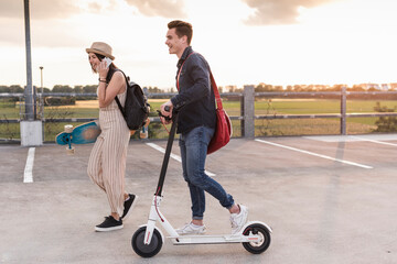 Happy young couple with cell phone, longboard and electric scooter on parking deck