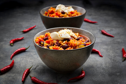 Red chili peppers and two bowls of vegetarian chili with red lentils, kidney beans, tomatoes, carrots, celery and sour cream