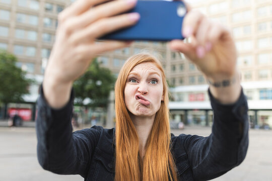 Portrait of redheaded young woman pouting mouth while taking selfie with smartphone, Berlin, Germany