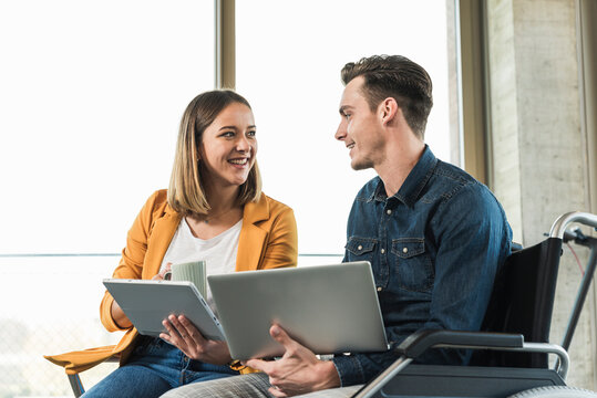 Young businessman with laptop in wheelchair and businesswoman with tablet in office