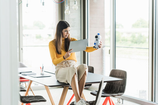 Young businesswoman sitting on table in office holding robot and tablet
