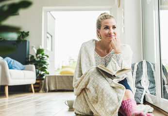 Woman wrapped in a blanket sitting at the window at home reading a book