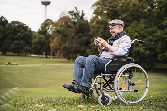 Happy senior man sitting in wheelchair on a meadow using smartphone