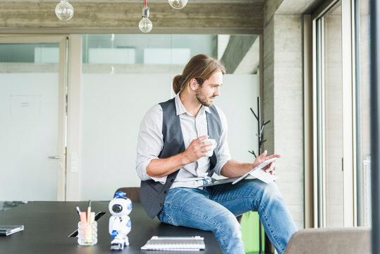 Young businessman sitting on table in office with tablet and cup of coffee