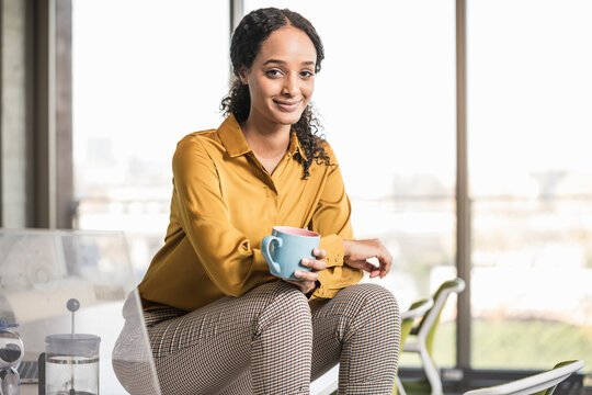 Portrait of smiling young businesswoman sitting on desk in office having a coffee break - Powered by Adobe