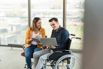 Happy young businessman with laptop in wheelchair and businesswoman with tablet in office