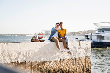 Happy young couple sitting on pier at the waterfront taking a selfie, Lisbon, Portugal