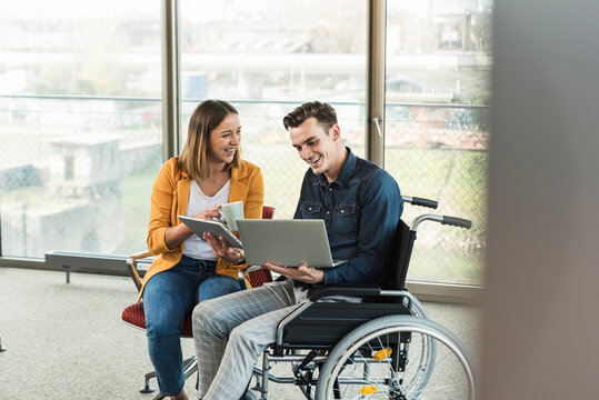 Happy young businessman with laptop in wheelchair and businesswoman with tablet in office