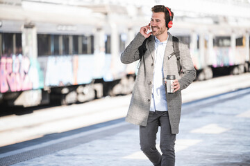 Happy young businessman with coffee mug and headphones walking at the train station