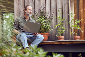 Thoughtful bearded man with laptop sitting outside tiny house
