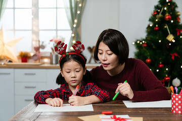 asian mother helping her young daughter making wish list of presents for Christmas at table in a bright room with Christmas decoration