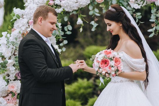 A young groom in a black suit puts a gold ring on the finger of a bride in a white dress on the background of an arch of flowers at the ceremony. Wedding portrait, photo of newlyweds.