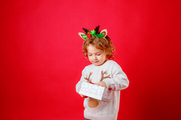 a little girl in deer horns holds a gift box on a red background, christmas, new year