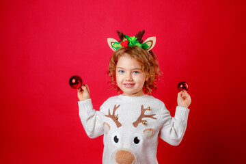 a little girl in deer horns holds Christmas tree toys on a red background, Christmas, new year