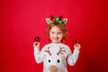 a little girl in deer horns holds Christmas tree toys on a red background, Christmas, new year