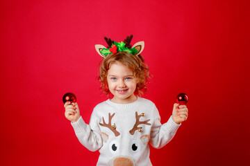 a little girl in deer horns holds Christmas tree toys on a red background, Christmas, new year