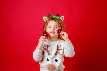 a little girl in deer horns holds Christmas tree toys on a red background, Christmas, new year