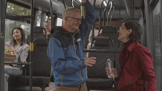 Medium side-view of short-haired Caucasian woman talking to senior man, standing in busy public transport, laughing