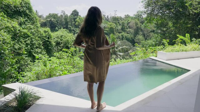 A young girl with long hair approaches the pool, throws off her robe and climbs from a modern white pool against a tropical landscape with green palm trees. A girl on vacation in a luxury spa hotel.