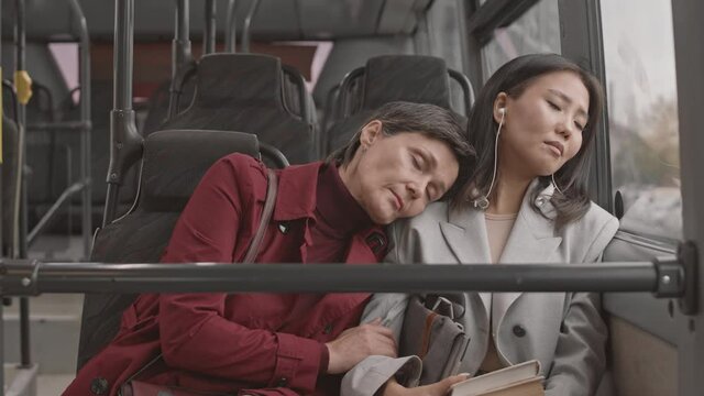 Waist-up of grey-haired Caucasian woman wearing red trench coat, sleeping on shoulder of young female Asian companion who also napping, then waking up and apologizing for it, sitting in bus