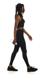 African american woman in black sports clothes is walking with bottle of water. Full length, side view, isolated.