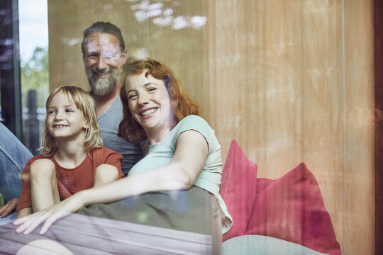 Smiling parents with daughter relaxing on bed at home seen through window