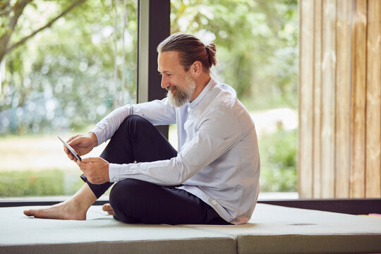 Smiling bearded mature man using digital tablet while sitting on bed by window at home