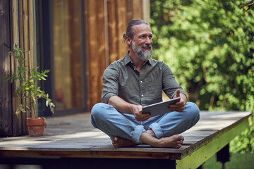 Bearded man with digital tablet contemplating while sitting outside tiny house