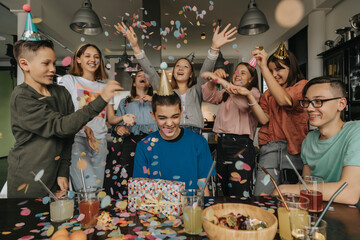 Cheerful friends throwing confetti on birthday boy sitting with gift at dining table