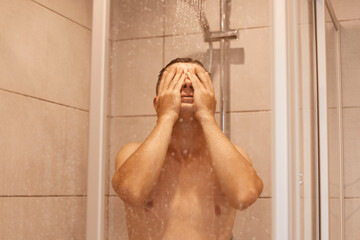 Portrait of young brunette relaxed man taking shower, standing under falling water drops, washing body and face in bathroom at home, bodycare and everyday hygiene routine.