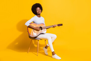 Full size profile portrait of pretty positive dark skin lady sit playing guitar isolated on yellow...