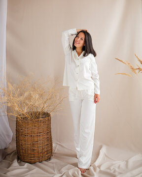 Full length portrait of happy beautiful young brunette woman wearing elegant white silk pajama set standing barefoot and smiling in cozy beige studio