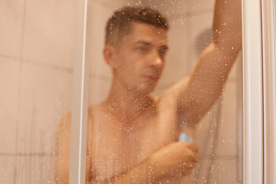 Indoor shot of young adult man washing his underarm with soap, looking at camera, handsome guy with dark hair taking shower, hygiene procedures after working day.