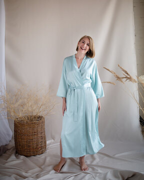 Full length portrait of beautiful young woman wearing elegant long turquoise silk robe sitting on stool barefoot