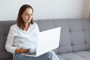 Portrait dark haired female wearing white casual style shirt and jeans, sitting on sofa in living room, looking at laptop display, having problems with eyesight.