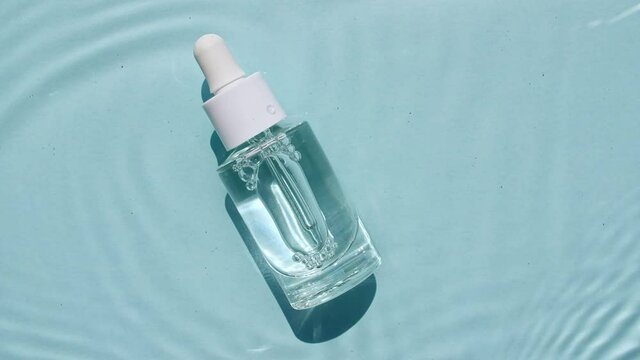 Glass cosmetic bottle with pipette lies on the surface of the water. Top view of water splash blue colored. Pure blue water with reflections sunlight. Sample of packaging for design, advertising.4k.