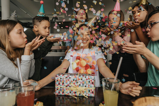 Happy friends throwing confetti on teenage girl sitting with birthday gift at dining table