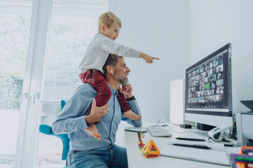 Mature businessman attending video conference while son sitting on his shoulder pointing on computer screen at home office