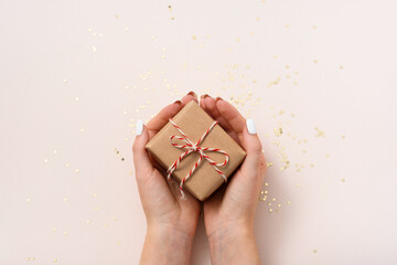 Female hands hold gift box in craft paper with white-red New Year's ribbon on a beige background
