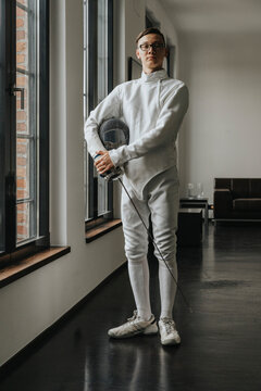 Young fencer with foil and mask standing by windows in loft at home