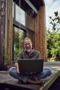 Smiling mature man with laptop sitting against tiny house