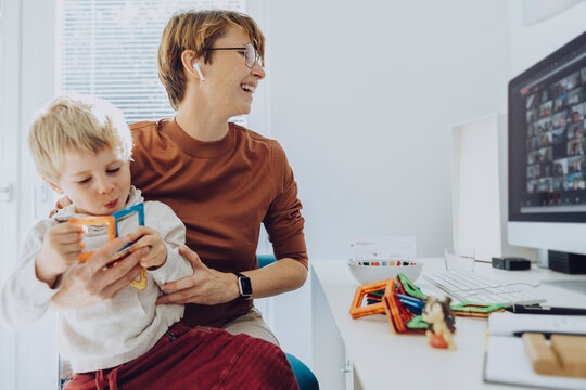 Mid adult businesswoman working while son sitting on her lap playing with toys at home office