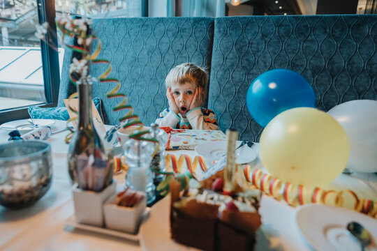 Surprised boy with hand on cheek sitting at table during birthday celebration in hotel