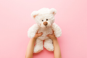 Smiling white teddy bear in baby girl hands on light pink background. Pastel color. Closeup. Point...