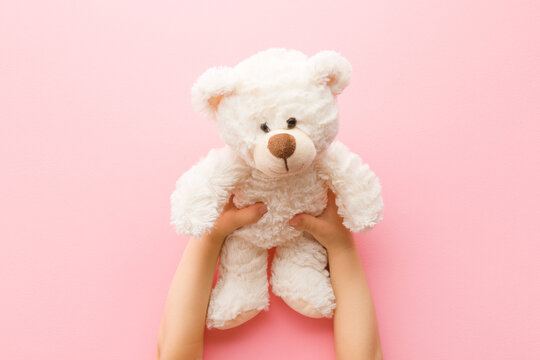 Naklejki Smiling white teddy bear in baby girl hands on light pink background. Pastel color. Closeup. Point of view shot. Kids best friend. Top down view.